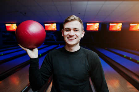 Cheerful young man holding a bowling ball and smiling at camera while standing against bowling alleys with ultraviolet light.の写真素材