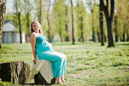 Happy pregnant woman dressed in a turquoise dress sitting on a stump at parkの写真素材