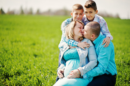 Happy pregnant family with two sons, dressed in a turquoise clothes, spending time together at green grass at fieldの写真素材