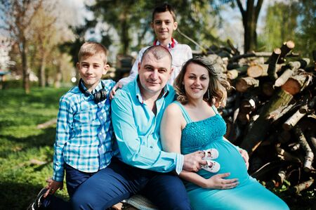 Happy pregnant family with two sons, dressed in a turquoise clothes sitting on a stump at park