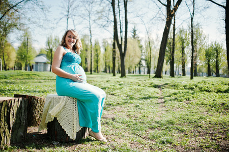 Happy pregnant woman dressed in a turquoise dress sitting on a stump at parkの写真素材