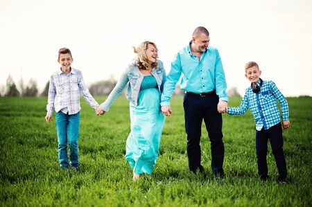 Happy pregnant family with two sons, dressed in a turquoise clothes, spending time together at green grass at fieldの写真素材