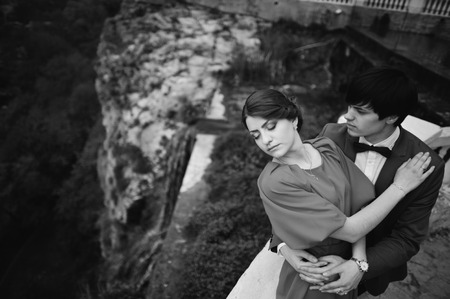 Couple hugging in love background fantastic cliff with waterfall. Stylish man at velvet jacket and girl in red dress in love together. Black and white photoの写真素材
