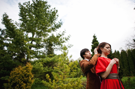 Couple hugging in love at beautiful landscape of trees and meadows. Stylish man at velvet jacket and girl in red dress in love togetherの写真素材