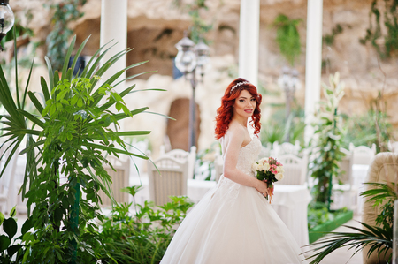 Charming red-haired bride model with wedding bouquet at hand posed at great wedding hall with caves and vegetation indoorの写真素材