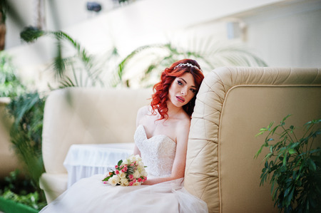 Portrait of charming red-haired bride model with wedding bouquet at hand sitting on the couch at the table on great wedding hall indoorの写真素材