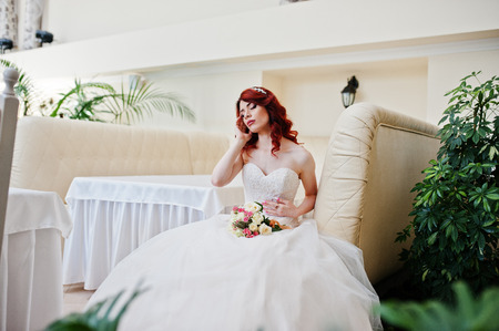 Portrait of charming red-haired bride model with wedding bouquet at hand sitting on the couch at the table on great wedding hall indoorの写真素材