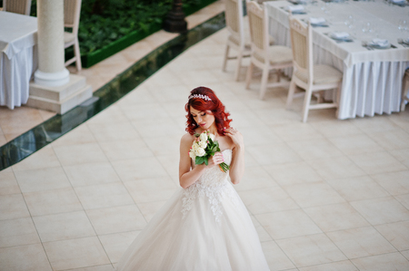 Charming red-haired bride with wedding bouquet at hand posed against the  great wedding hall with tables and chairs indoorの写真素材