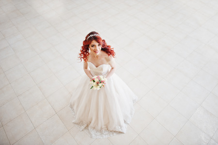 Top view of charming red-haired bride with wedding bouquet at hand posed against wgite tile on the great wedding hallの写真素材