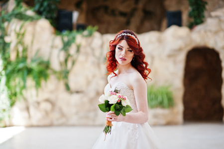 Close up portrait of charming red-haired bride with wedding bouquet at hand posed at great wedding hall with caves and vegetation indoorの写真素材