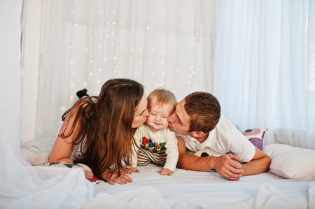 Mother and father with baby boy lying on white bed with garland