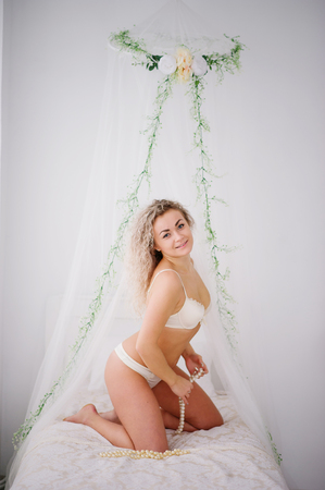 Curly blonde girl in underwear sitting on bed indoor white room.の写真素材