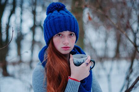 Portrait of young red hair girl with freckles wearing at blue knitted wool hat and scarf with cup of tea in winter day.の写真素材