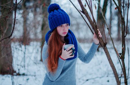 Portrait of young red hair girl with freckles wearing at blue knitted wool hat and scarf with cup of tea in winter day.の写真素材