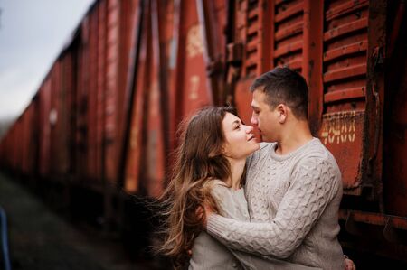 Young couple wearing on tied warm sweaters hugging in love at railway stations background freight cars.の写真素材