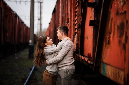 Young couple wearing on tied warm sweaters hugging in love at railway stations background freight...