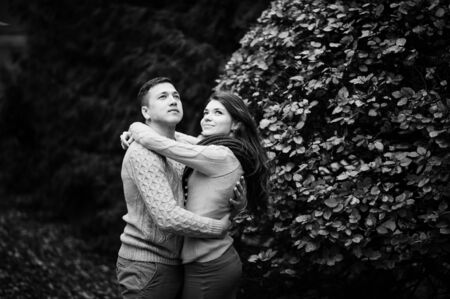 Young couple wearing on tied warm sweaters hugging in love at city in autumn background yellow bushes and trees.の写真素材