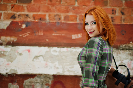 An outdoor portrait of a young pretty girl with red hair wearing checkered dress with girly backpacks standing on the brick wall background.の写真素材