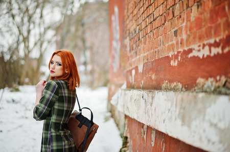 An outdoor portrait of a young pretty girl with red hair wearing checkered dress with girly backpacks standing on the brick wall background in winter day.の写真素材
