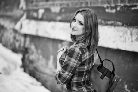 An outdoor portrait of a young pretty girl with red hair wearing checkered dress with girly backpacks standing on the brick wall background in winter day. Black and white photoの写真素材