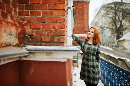 An outdoor portrait of a young pretty girl with red hair wearing checkered dress with girly backpacks standing on the brick wall background in winter day.の写真素材