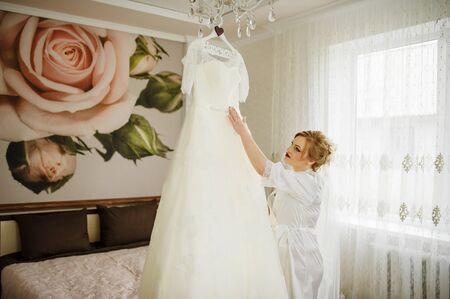 Blonde bride in a bathrobe looking at wedding dress at her room.の写真素材