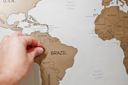 Scratch travel map of the world. Hand of man erase Brazil with coin.