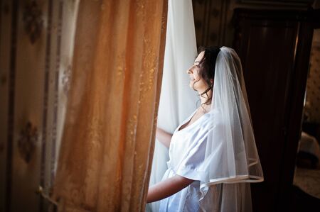 Brunette bride looking at window on hotel room at morning wedding day.の写真素材