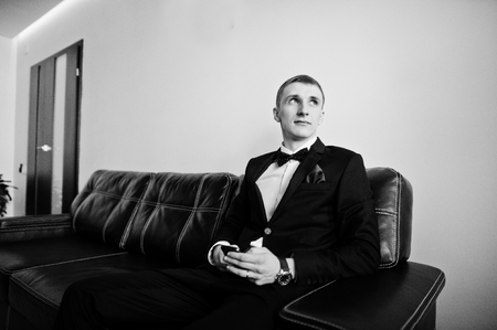 Young stylish groom sitting on leather sofa. Black and white photoの写真素材