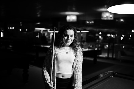 Young curly girl posed near billiard table. Sexy model at black mini skirt play russian snooker....