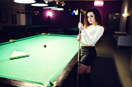 Young curly girl posed near billiard table. Sexy model at black mini skirt play russian snooker. Play game and fun concept.の写真素材