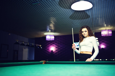 Young curly girl posed near billiard table. Sexy model at black mini mini skirt play russian snooker. Play game and fun concept.の写真素材