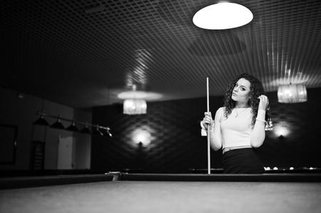 Young curly girl posed near billiard table. Sexy model at black mini skirt play russian snooker. Play game and fun concept.の写真素材