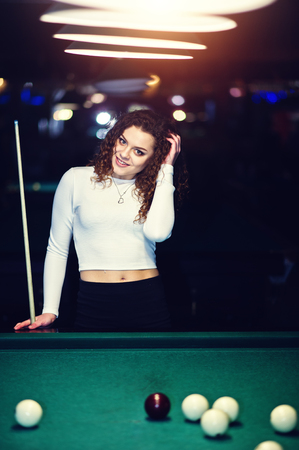 Young curly girl posed near billiard table. Sexy model at black mini skirt play russian snooker. Play game and fun concept.の写真素材