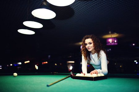 Young curly girl posed near billiard table. Sexy model at black mini skirt play russian snooker....