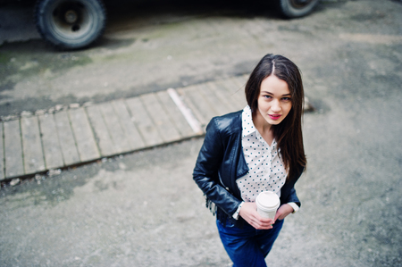 Portrait of stylish young girl wear on leather jacket and ripped jeans with cup of coffee. Street fashion model style.の写真素材