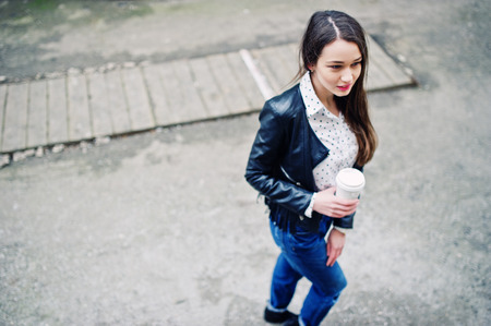 Portrait of stylish young girl wear on leather jacket and ripped jeans with cup of coffee. Street fashion model style.の写真素材