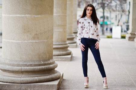 Young stylish brunette girl on shirt, pants and high heels shoes, posed near stone columns. Street fashion model concept.の写真素材
