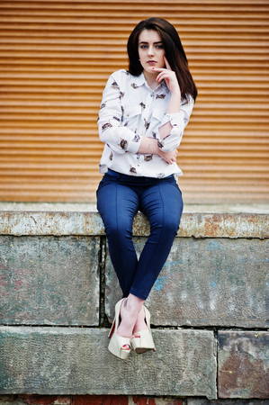 Young stylish brunette girl on shirt, pants and high heels shoes, sitting and posed background orange shutter. Street fashion model concept.の写真素材