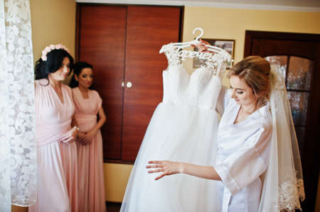 Bride on silk robe looking on her wedding dress against bridesmaids at room on morning day.の写真素材