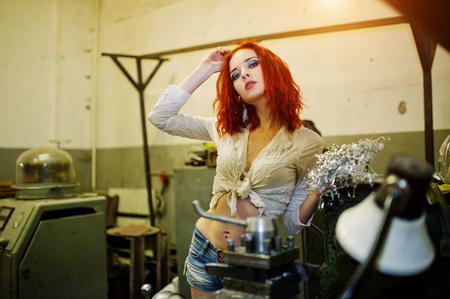 Red haired girl wear on short denim shorts and white blouse posed at industrial machine at the factory.の写真素材