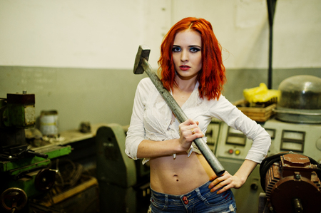Red haired girl wear on short denim shorts and white blouse with hammer at hands posed at industrial machine at the factory.の写真素材
