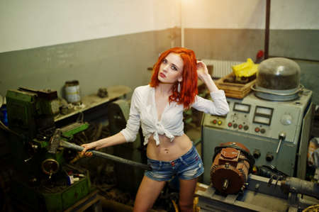 Red haired girl wear on short denim shorts and white blouse with hammer at hands posed at industrial machine at the factory.の写真素材