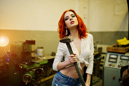 Red haired girl wear on short denim shorts and white blouse with hammer at hands posed at industrial machine at the factory.の写真素材