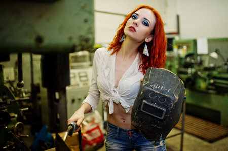 Red haired girl wear on short denim shorts and white blouse with welding mask at hands posed at industrial machine at the factory.の写真素材