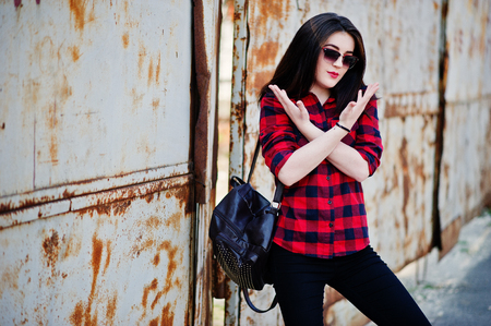 Fashion portrait girl with red lips wearing a red checkered shirt, sunglasses and backpack background rusty fence.の写真素材