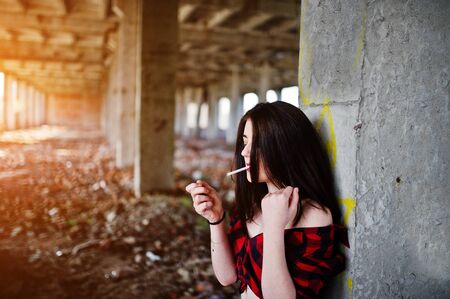 Portrait smoking girl with red lips wearing a red checkered shirt with bare shoulders posed sexy background abadoned place.の写真素材