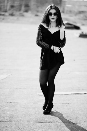 Portrait brunette girl with red lips wearing a black dress, tights and shoes on heels posed on the roof. Street fashion model.の写真素材