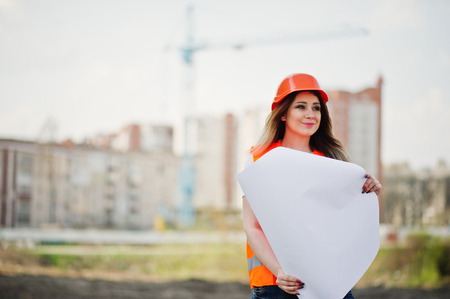 Engineer builder woman in uniform waistcoat and orange protective helmet hold business paper against new buildings with crane.の写真素材