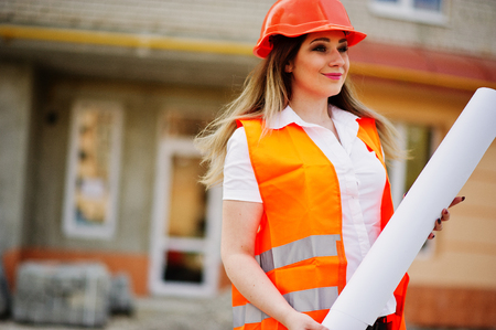 Engineer builder woman in uniform waistcoat and orange protective helmet hold business paper against new building. Property living block theme.の写真素材